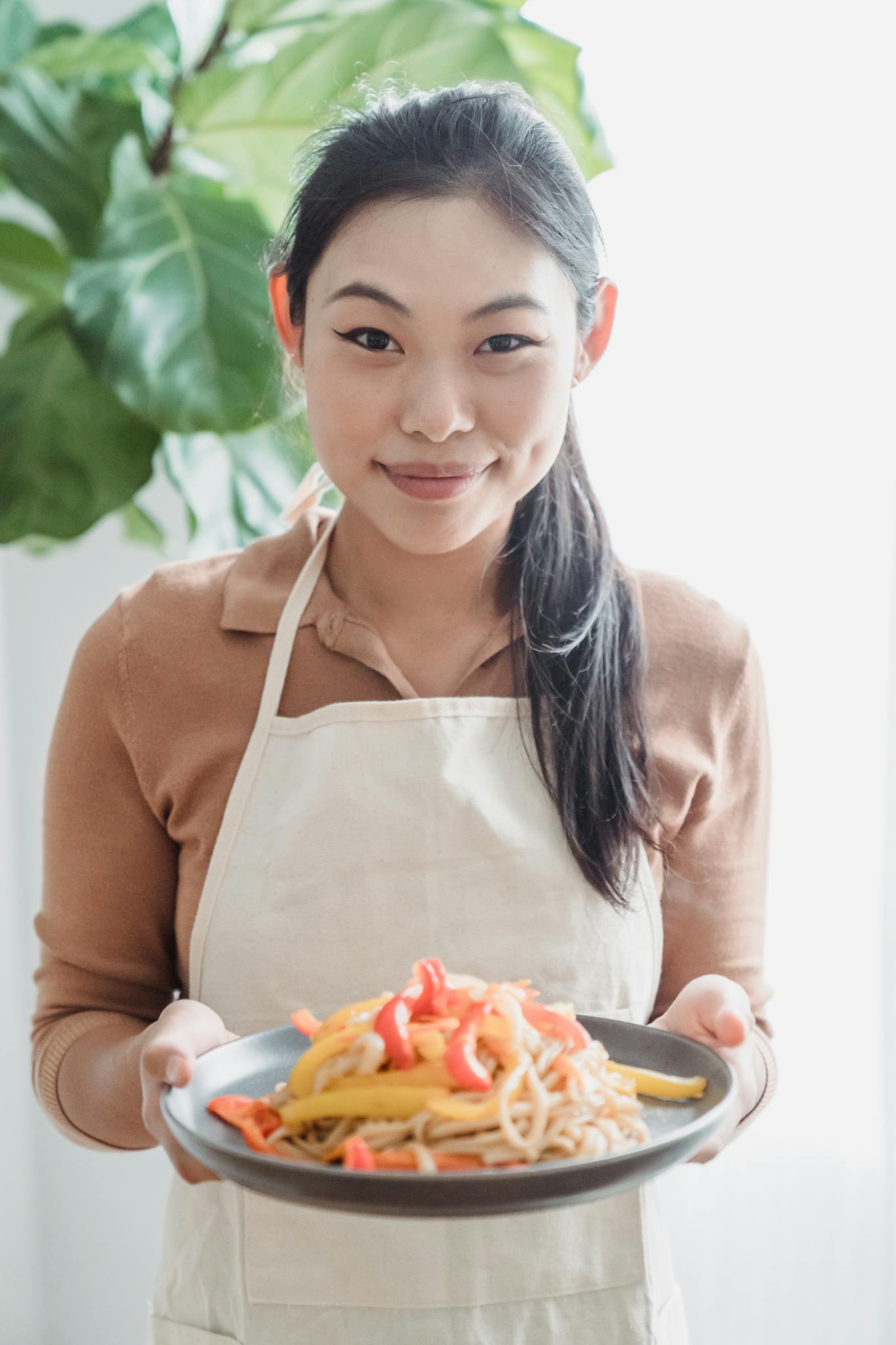 Smiling woman presenting a vibrant plate of pasta with vegetables indoors.